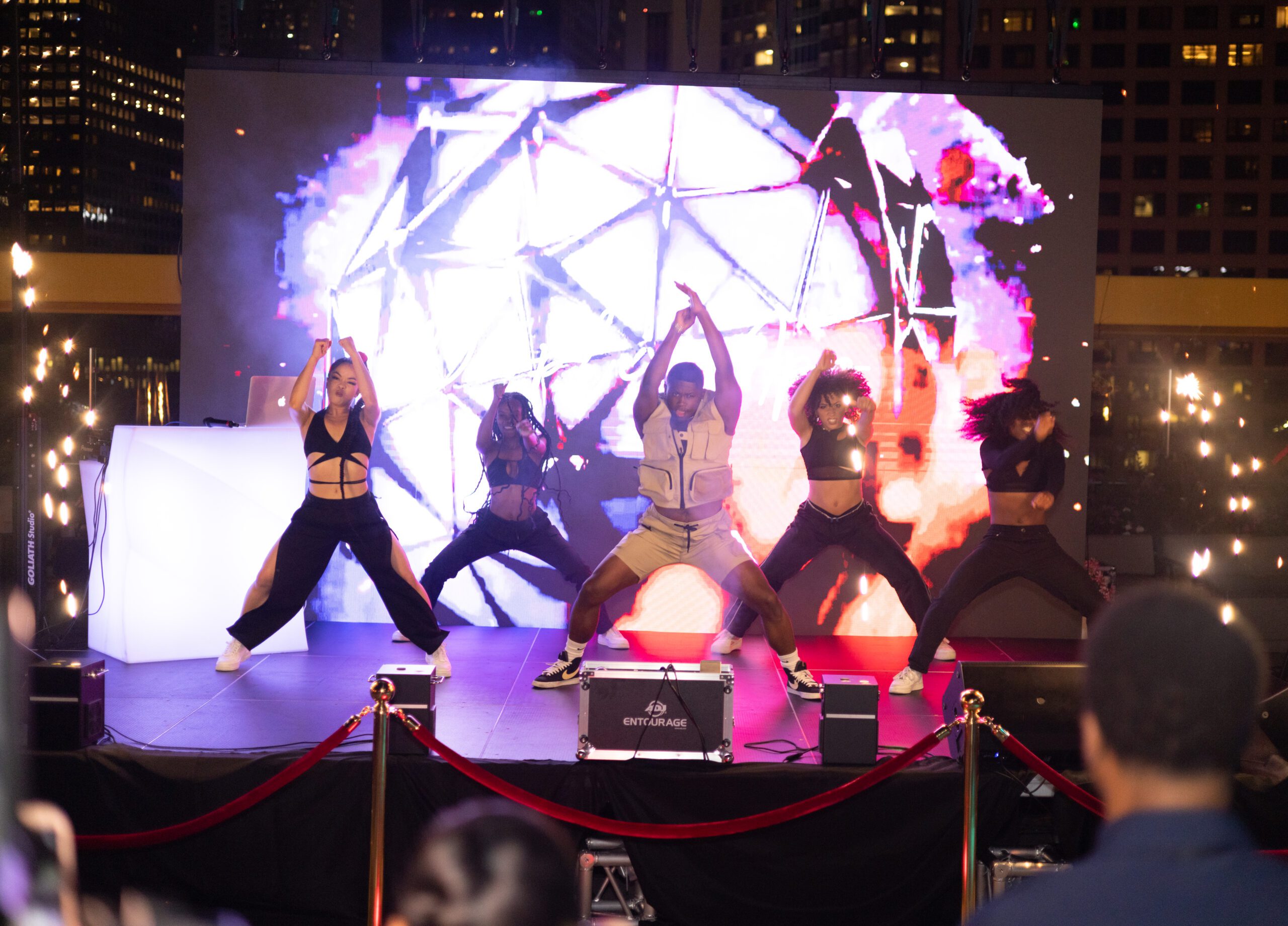 a group of dancers performing energetically on a brightly lit stage with a colorful, dynamic background