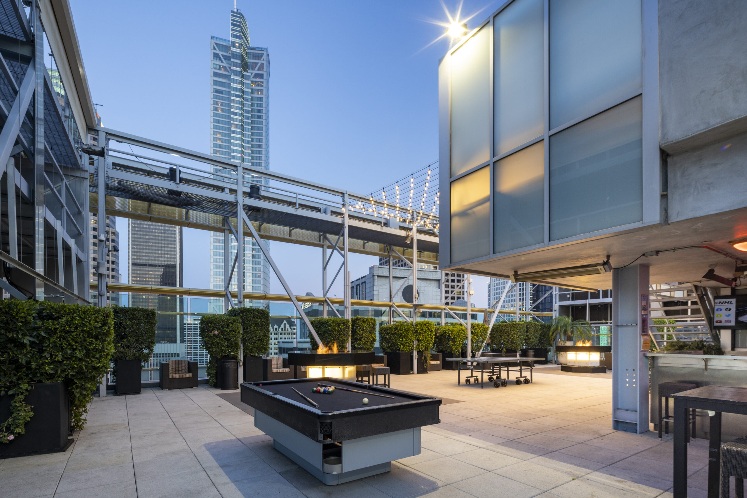 a modern outdoor rooftop lounge with a pool table, seating areas, greenery, and a city skyline view.